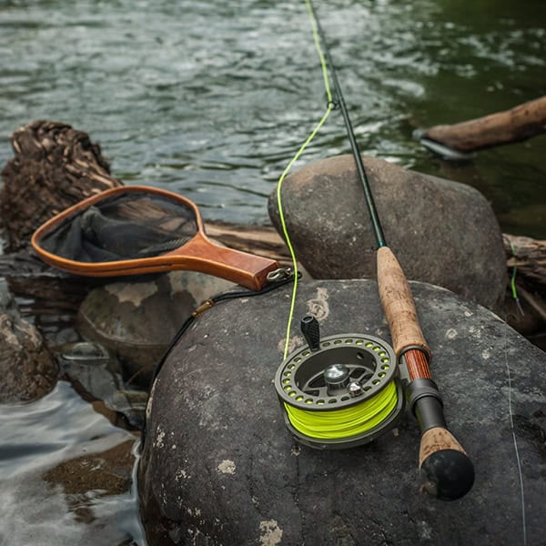 A rod, reel, and net laying on large river rocks on the banks of a river.