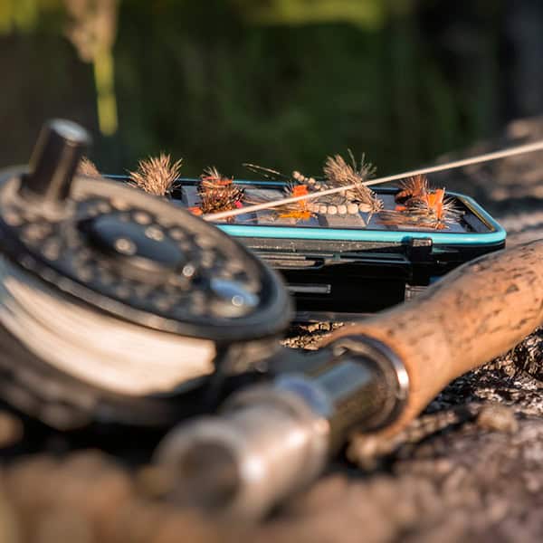 A reel and a box of fly fishing flies lay in the gravel.