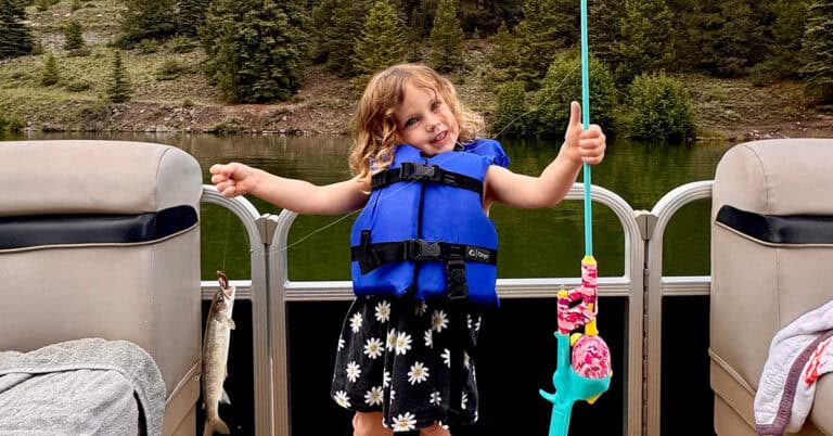 a little girl in a blue life vest playfully shows off her fishing pole and recent catch of a trout.