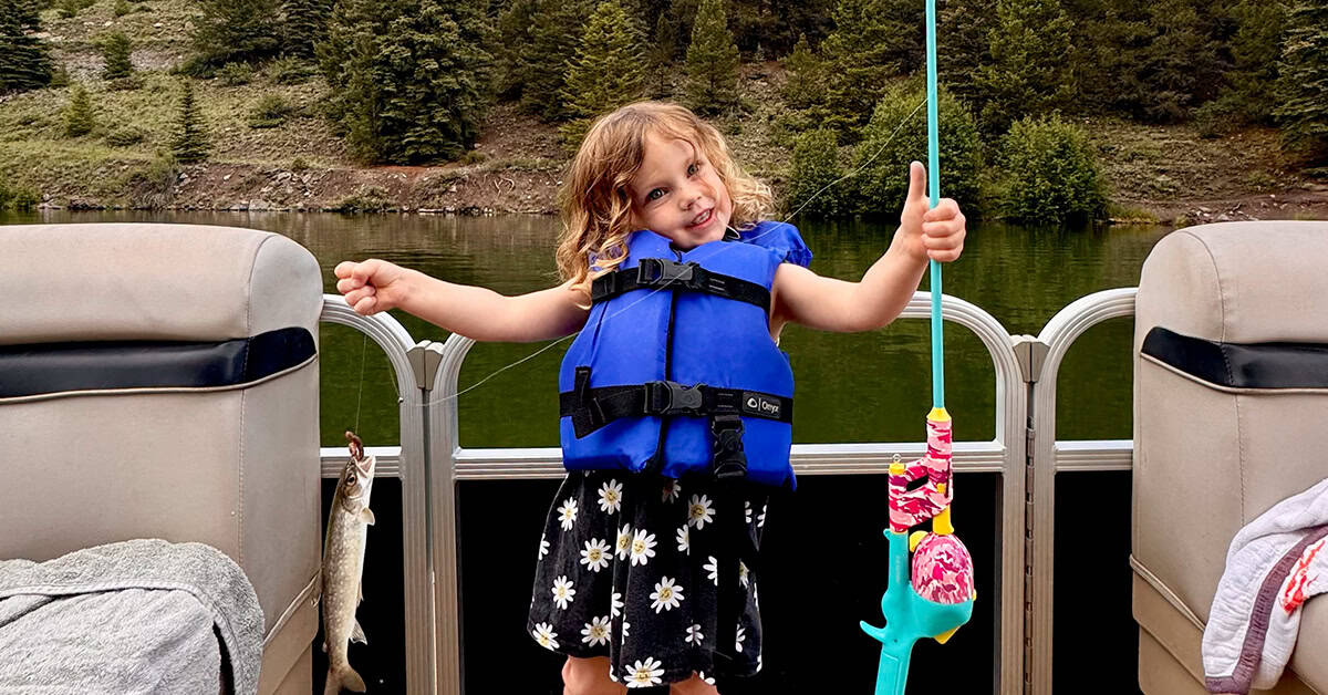 a little girl in a blue life vest playfully shows off her fishing pole and recent catch of a trout.