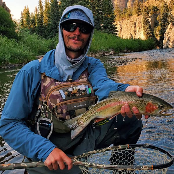 Joe Miller kneeling in a mountain stream with a beautiful rainbow trout wearing a decked out fly fishing vest and net in front of a golden bluff raising out of the river in the background.