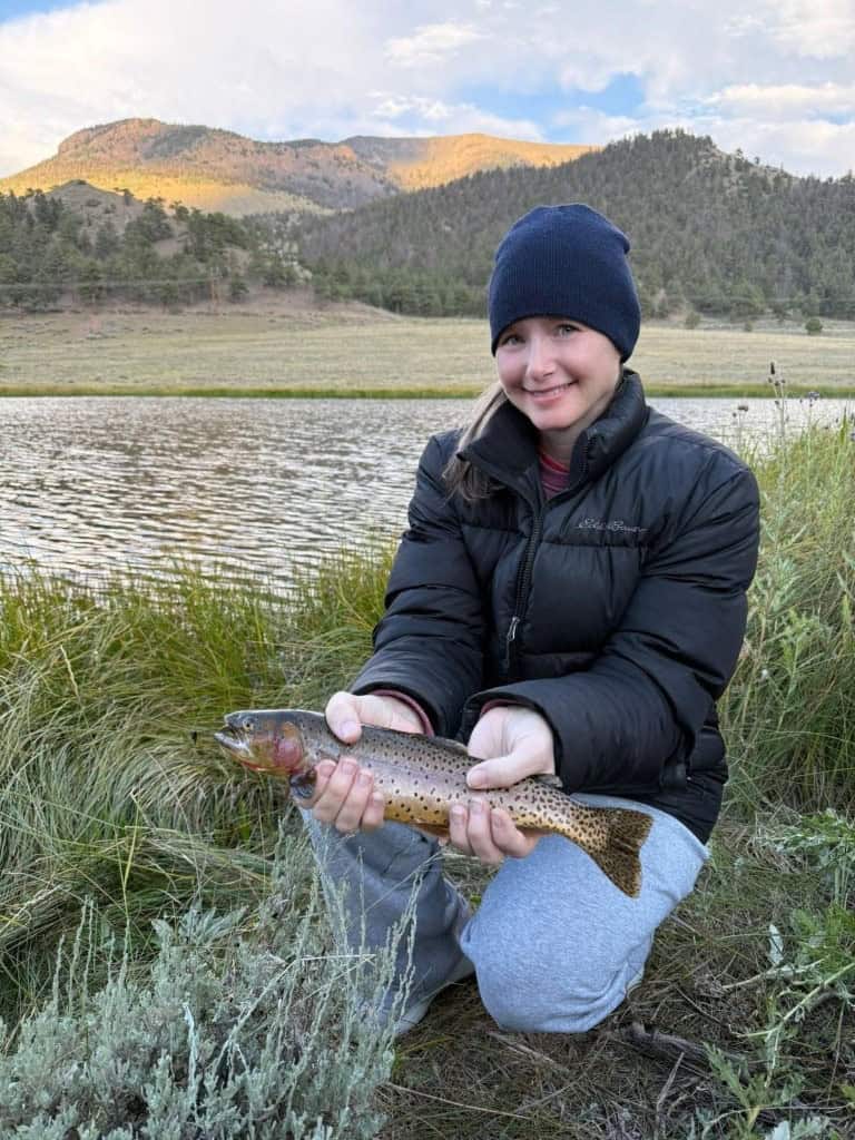 A quiet smile shows the pride of the catch for this young lady kneeling beside a large pond with a beautiful trout.
