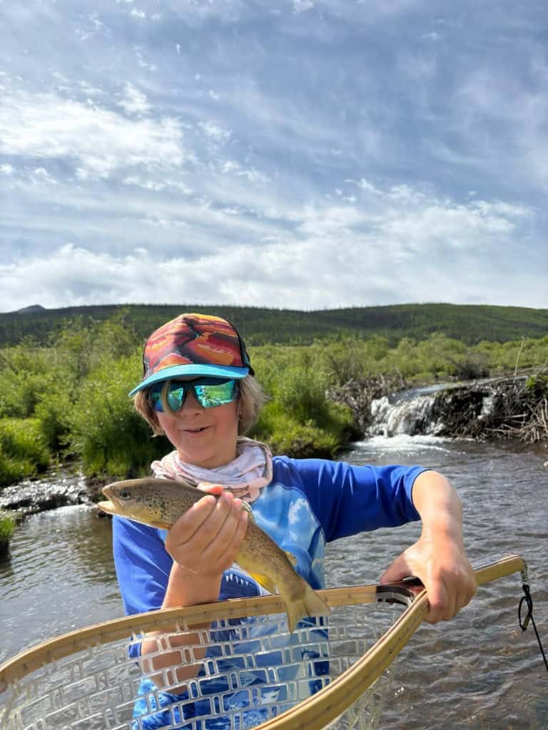 A young angler pulls a trout from a net with a look of wonder with a creek and willows behind.