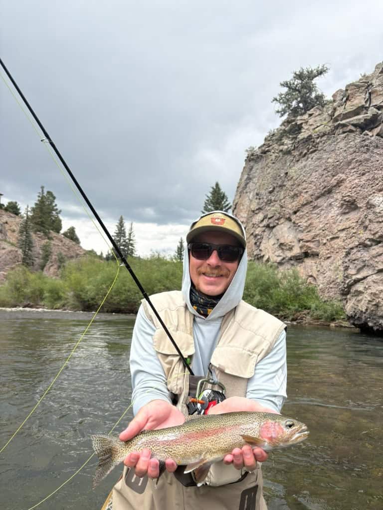 A beautiful rainbow trout is held in two hands by an angler with a big grin on his face from the river.
