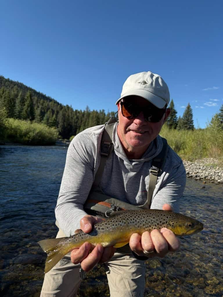 A man in a ball cap displays his nice trout catch while standing in a river surrounded by mountains on a beautiful sunny day.