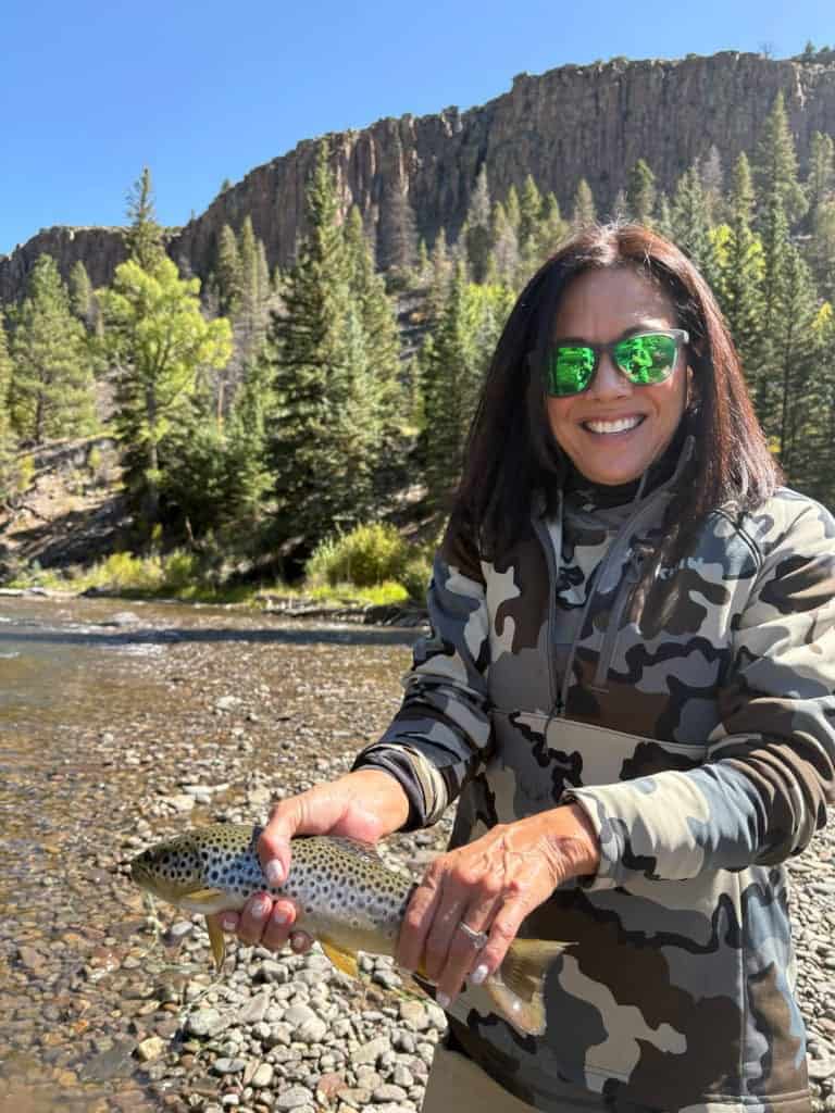 A woman with dark hair and sunglasses in a camo pullover smiles with her spotted trout catch beside the river with a rock wall towering above.