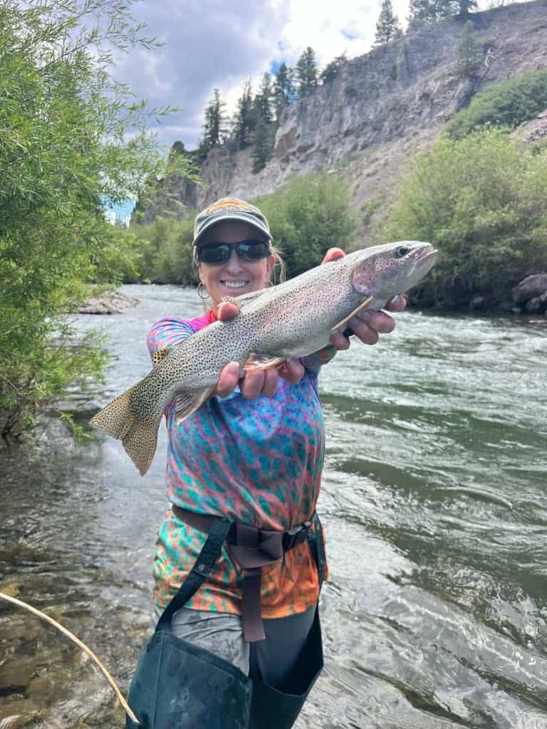 Woman holding a large fish in a river with trees and cliffs in the background.
