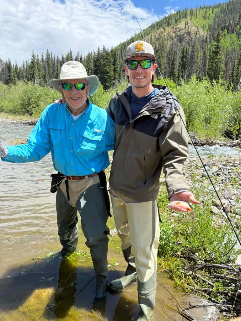 Fishing at a river with two anglers in outdoor gear, surrounded by lush trees and mountains.