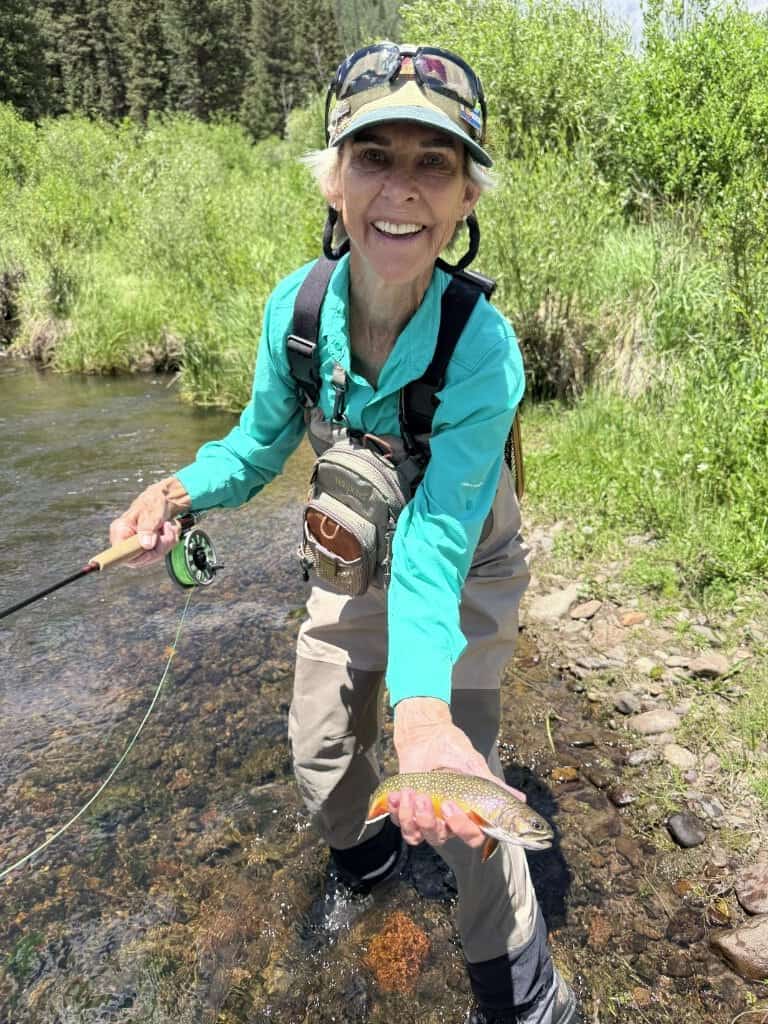 A lady angler proudly shows her brookie while standing at river's edge and holding her fly rod.