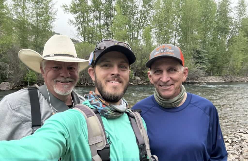 Guide, Mitchell Murphy, stands between two happy customers beside a river and cottonwood trees.