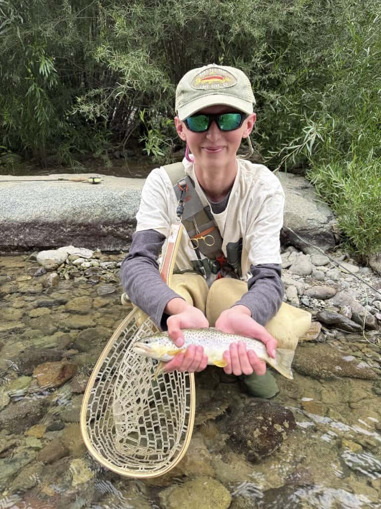 An angler wearing fly fishing gear smiles and holds a trout with yellow fins.