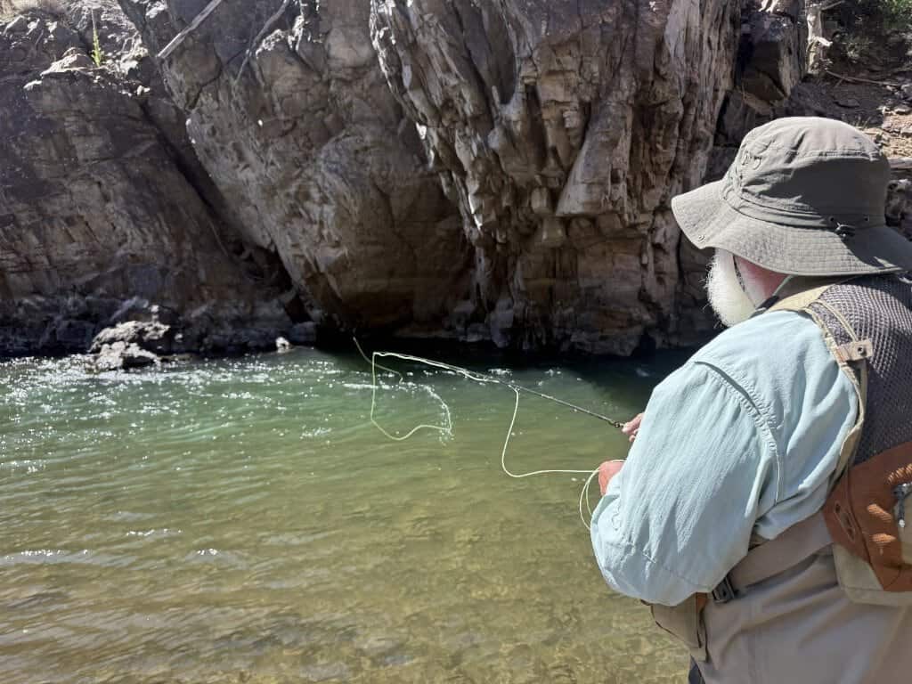 A gentleman with a white beard and bucket hat places his fly line in the perfect spot in the river below a cliff.