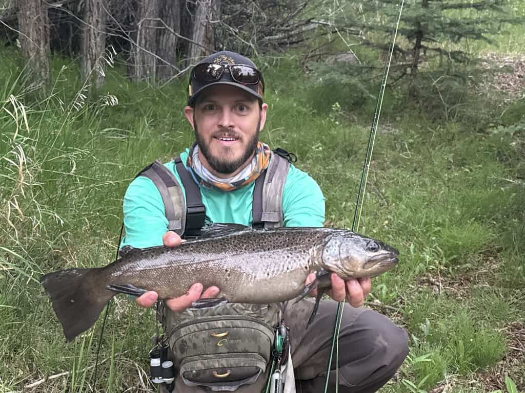 Mitchell Murphy holds an astounding trout caught on a fly rod while kneeling in the grass.
