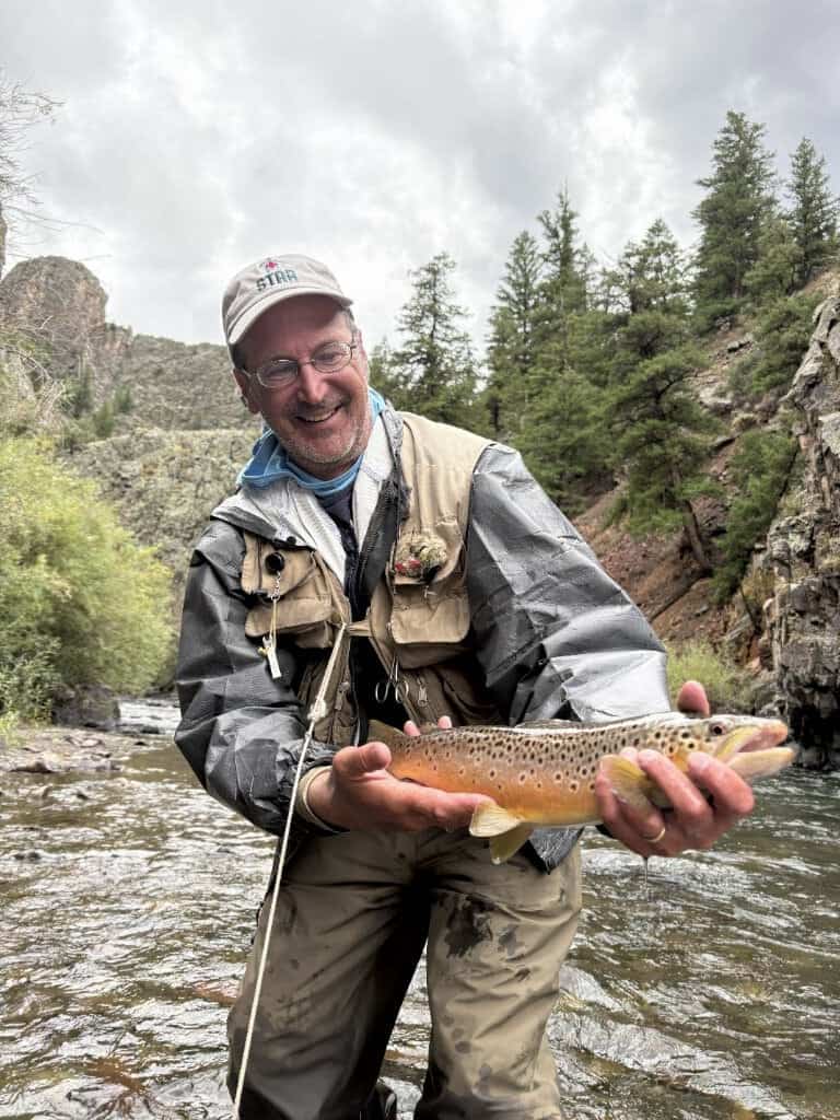 A nice trout is held by a man wearing fly fishing vest and pants in a mountain stream beside sagebrush hills and pines.